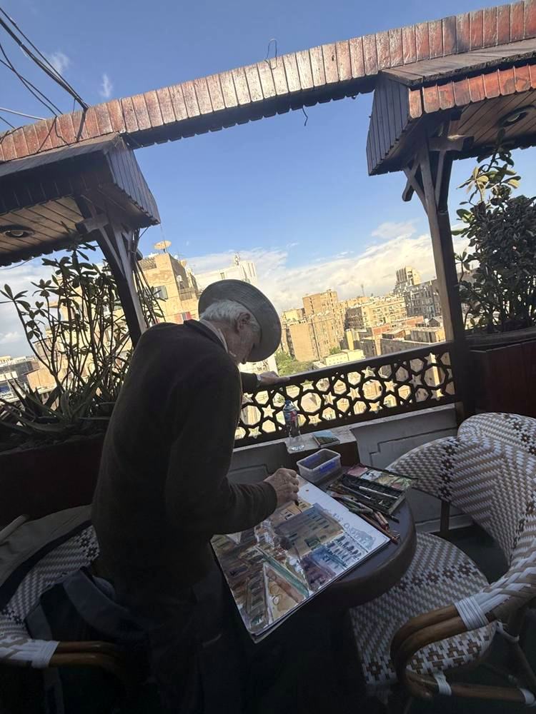 An older man in a hat painting the Downtown view from the rooftop terrace
